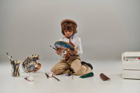 young painter mixing paint on palette, boy in beret surrounded by paint brushes and colorful tubes