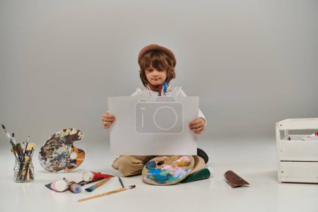 young artist surrounded by vibrant colors and tools for painting, boy looking at blank paper