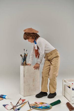 elegante artista se apoya en un bloque blanco con pinceles, niño en boina y bufanda en el estudio