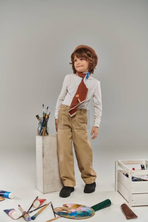 stylish artist standing near white block with paint brushes, boy in beret and scarf in studio