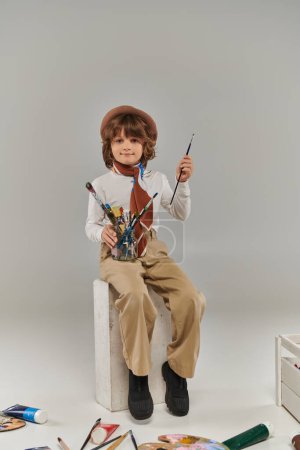 artiste élégant assis sur bloc blanc avec pinceaux, garçon en béret et écharpe en studio