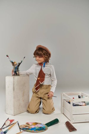 young artist sitting near white block with paint brushes, stylish boy in beret and scarf in studio