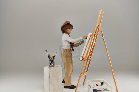 junger Künstler beim Malen im Stehen mit Palette neben hölzerner Staffelei, stylischer Junge mit Baskenmütze und Schal