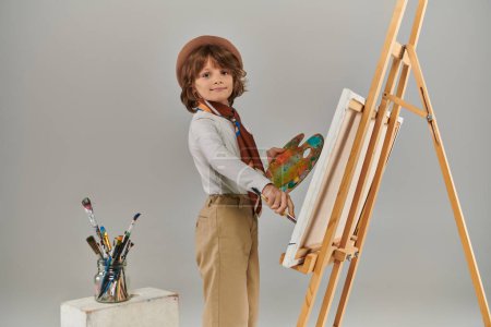 Foto de Niño feliz explora su potencial creativo, artista en boina con colorida paleta de pintura cerca del caballete - Imagen libre de derechos