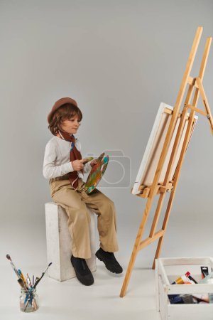 niño feliz explora su potencial creativo, artista en boina con paleta mirando caballete con lienzo