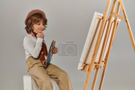 fröhlicher junger Künstler erkundet sein kreatives Potenzial, Junge in Baskenmütze blickt auf Staffelei mit Leinwand