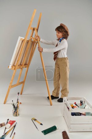 Téléchargez les photos : Jeune artiste ajustant son chevalet, entouré de ses outils et peinture colorée, garçon en béret et écharpe - en image libre de droit