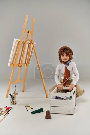 Téléchargez les photos : Heureux jeune artiste choisir la peinture dans la boîte en bois, garçon dans le béret assis près du chevalet et pinceaux - en image libre de droit