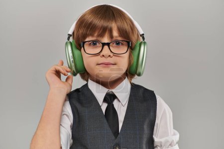 stylischer Junge in Brille und eleganter Kleidung mit Weste, der über grüne Kopfhörer Musik hört