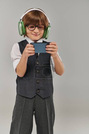 stylischer Junge mit Brille und eleganter Kleidung, der über Kopfhörer Musik hört und Handyspiele spielt