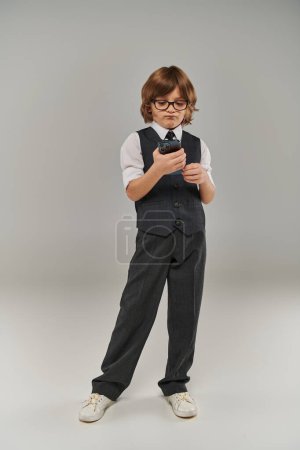 displeased boy in glasses and elegant attire  playing mobile game on grey, holding smartphone