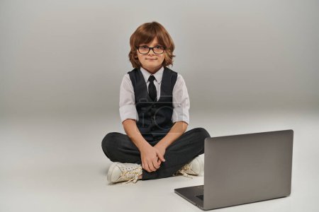 niño en traje elegante con chaleco y pantalones sentados cerca de la computadora portátil en gris, futuro hombre de negocios