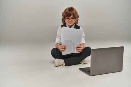Junge in eleganter Kleidung mit Weste sitzt neben Laptop und schaut auf Papiere, zukünftiger Geschäftsmann