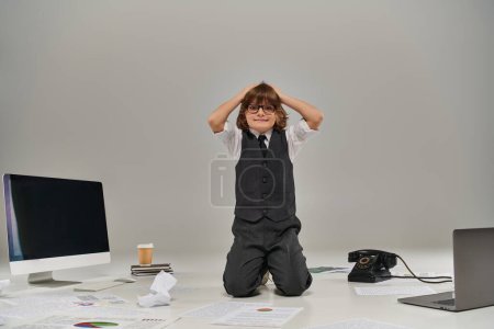 stressed boy in glasses surrounded by papers and office supplies on grey, future businessman