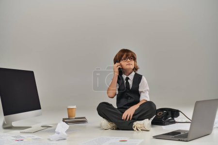 Junge in Brille und festlicher Kleidung, der mit dem Smartphone spricht und von Bürogeräten umgeben sitzt