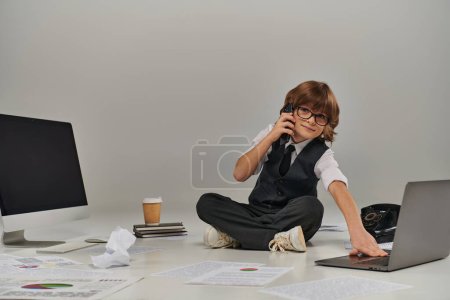 Kind in Brille und offizieller Kleidung spricht auf Smartphone und sitzt umgeben von Bürogeräten