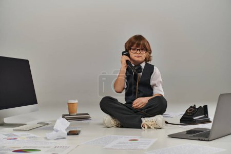Kind mit Brille und formeller Kleidung, das am Retro-Telefon spricht und von Bürogeräten umgeben sitzt