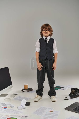 niño en gafas y ropa formal rodeado de equipos de oficina y dispositivos de pie en gris