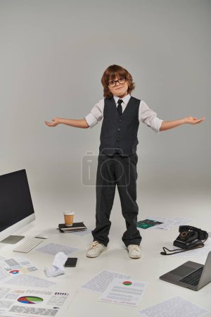 confused kid in glasses and formal wear surrounded by office equipment and devices standing on grey