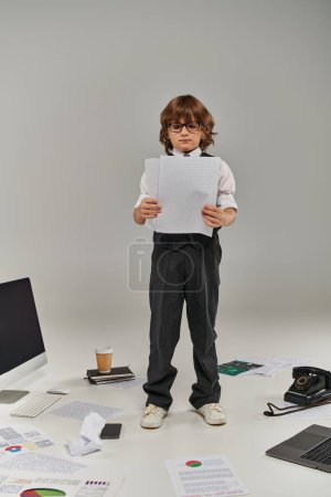 cute kid in glasses and formal wear surrounded by office equipment and devices standing with papers