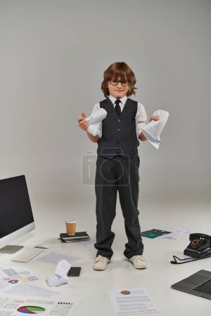 Netter Junge in Brille und formaler Kleidung, umgeben von Bürogeräten und Geräten, die mit Papieren stehen