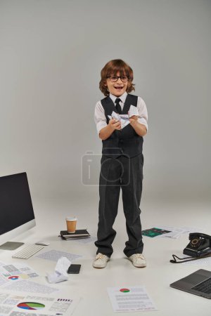 excited boy in glasses and formal wear surrounded by office equipment and devices holding papers