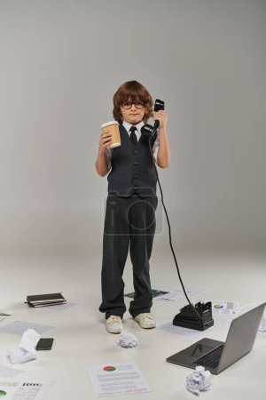 Netter Junge mit Brille und formaler Kleidung, der Papierbecher und Retro-Telefon während des Gesprächs hält, Multitasking