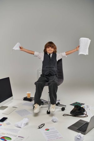 glücklicher Junge mit Papieren in den Händen, der auf einem Stuhl sitzt, umgeben von Büromaterial, zukünftiger Berufstätiger