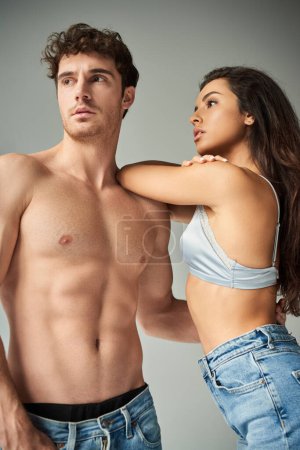 hübsche junge Frau in Satin-BH und Jeans umarmt hemdlosen Mann auf grauem Hintergrund, Zuneigung