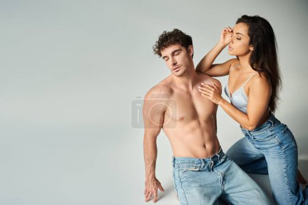 sensual joven en sujetador de seda y jeans posando con hombre sin camisa sobre fondo gris, cercanía