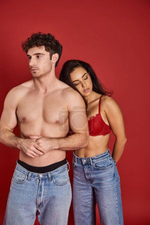 Photo for Sensual young woman in bra and denim jeans leaning on shirtless man on red background, affection - Royalty Free Image