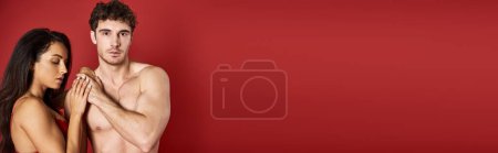 Photo for Muscular man looking at camera near gorgeous brunette woman in bra on red backdrop, banner - Royalty Free Image