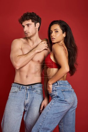 muskulöser und hemdloser Mann in Jeans, der mit einer hinreißenden brünetten Frau im BH vor rotem Hintergrund steht