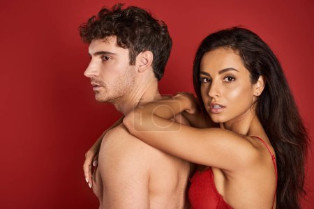 gorgeous and brunette woman in bra leaning on handsome shirtless boyfriend on red background