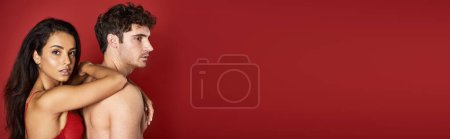 Photo for Gorgeous and brunette woman in bra leaning on handsome shirtless boyfriend on red background, banner - Royalty Free Image