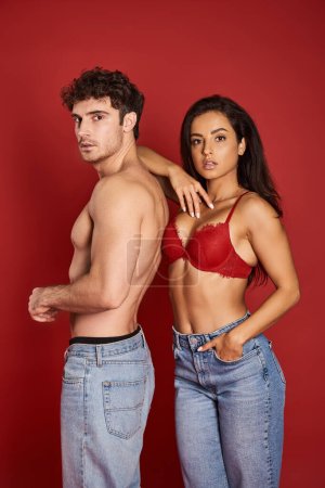 gorgeous young woman in bra  posing with hand in pocket of jeans near shirtless boyfriend on red