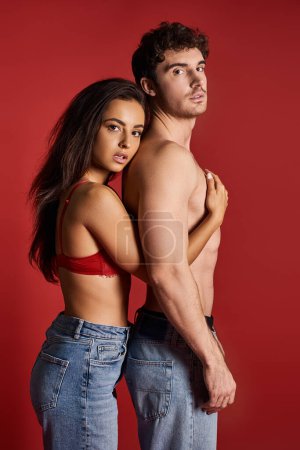 Photo for Brunette and charming young woman in lace bra and jeans embracing her muscular man on red background - Royalty Free Image