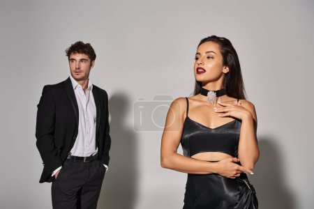 attractive woman posing in black dress near handsome man in formal wear on grey background