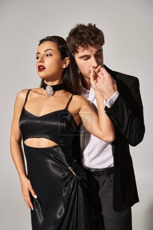 Photo for Handsome man in suit kissing hand of brunette young woman in black slip dress on grey background - Royalty Free Image