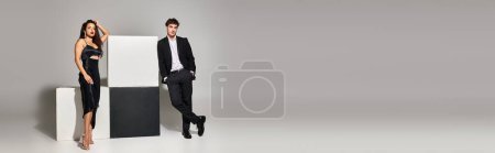 handsome man in suit posing with hands in pockets near cubes and gorgeous woman on grey, banner