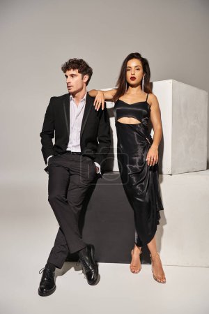 man in suit posing with hands in pockets and looking at gorgeous brunette woman on grey backdrop