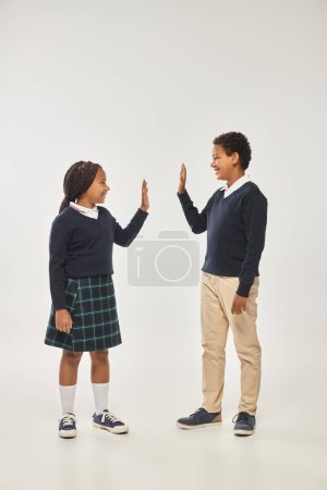 happy african american schoolkid in uniform giving high five to each other on grey background