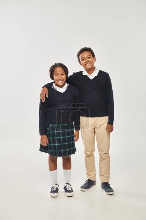 happy african american boy in school uniform hugging girl in plaid skirt while standing on grey