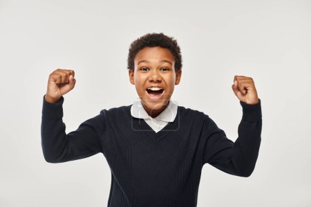 excited african american schoolboy in uniform rejoicing while looking at camera on grey backdrop Poster 692617474