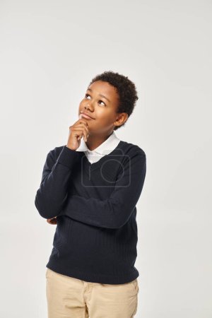 pensive african american boy in school uniform touching chin while thinking on grey backdrop