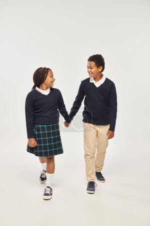happy preteen african american schoolchildren in uniform holding hands on grey background