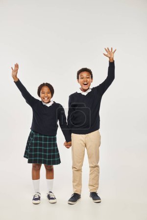 excited preteen african american schoolchildren in uniform holding hands on grey background