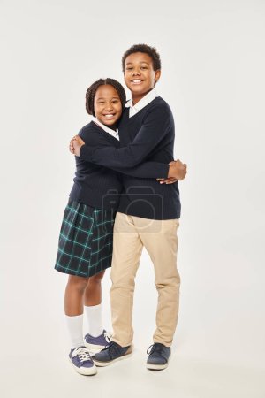cheerful preteen african american schoolchildren in uniform hugging each other on grey background