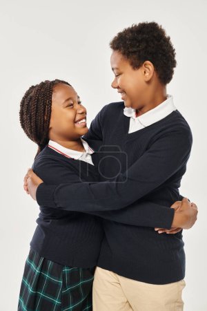 fröhliche preteen afrikanisch-amerikanische Kinder in Schuluniform umarmen einander auf grauem Hintergrund