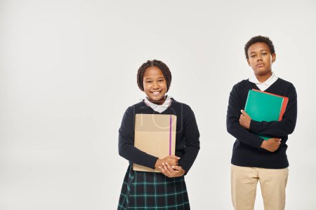 happy african american schoolkids in neat uniform holding textbooks and standing on grey backdrop
